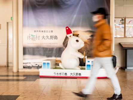 A blurred figure of a man in white trousers, light tan anorak and black baseball cap walks across a stone floor in front of a large white figurine of a rabbit.