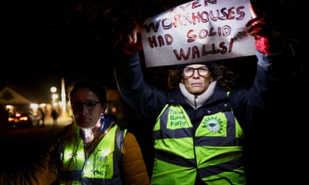 Two protesters at a vigil outside Manston wearing Green party high-vis vests, with one holding a sign saying: Even Workhouses Had Solid Walls