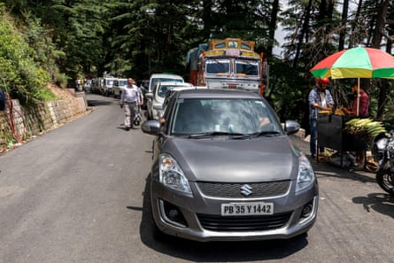 New-looking cars in a line bumper to bumper on a narrow road passing a stall selling sweetcorn.
