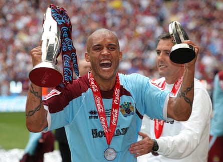 A man in a blue and burgundy football kit holding a silver football trophy in one hand and a small trophy in another hand