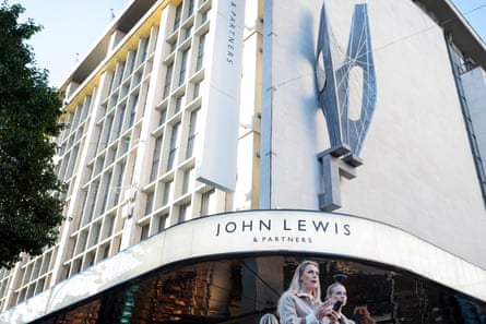 Frontage of the John Lewis store on Oxford Street, London, with an angular modernist sculpture above the doorway that curves around one corner. Two women walk in the foreground and the upper storeys’ white facade with recessed windows stretches along the street.