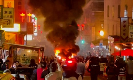Smoke billows from a car on fire as a crowd of people surround it.