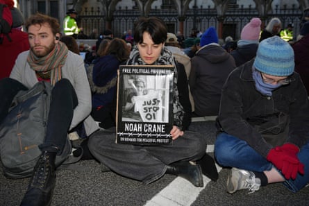 Three young protesters sit cross-legged in the road; one holds a placard with a picture of a jailed activist. Other activists sit in the background; many of them wear woolly hats.