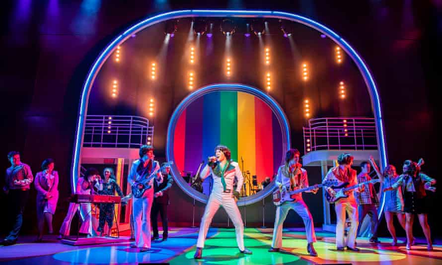 Cast members in white jumpsuits against rainbow-coloured backdrop