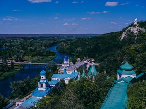 A large complex of buildings with blue onion domes on a hillside overlooking a river with a white monumental statue in the background