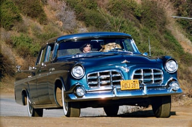 A woman drives a vintage blue car in California while a lion sits in the passenger seat