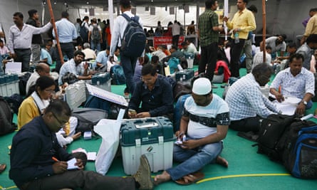 Dozens of people sitting in groups on the floor making notes and reading documents, with the machines in sealed boxes