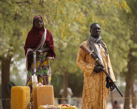 A man with a gun stands guard while a woman pumps water into jerry cans from a tap.