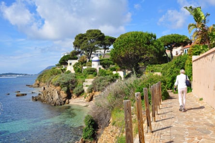 A person in white walks away from the camera, down a paved path towards trees, with sea off to the left