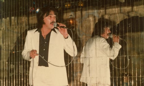 José Pinhal sings before a mirrored wall, in black shirt and white suit.