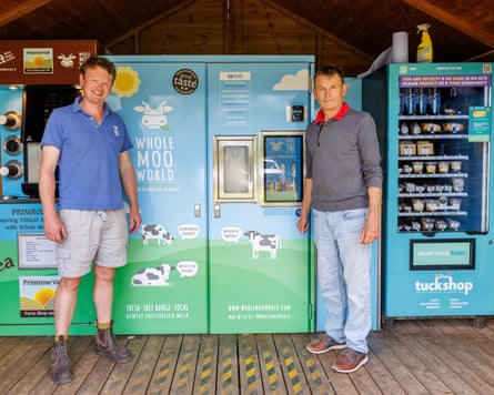 Mark and Sam stand in front of their blue and green vending machines, which are installed in a wooden hut.