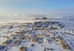 Aerial picture of abandoned buildings in a snowy landscape. The still-working coalmine is in the distance behind, belching smoke