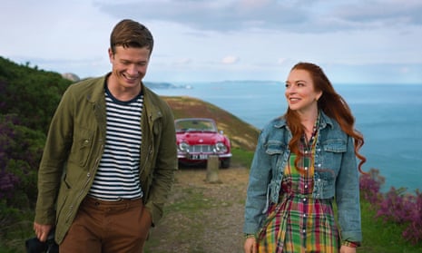 A color movie still of a white man and a white woman in spring clothes and jackets, smiling and walking along a grassy cliff alongside the ocean, with a red convertible behind them.