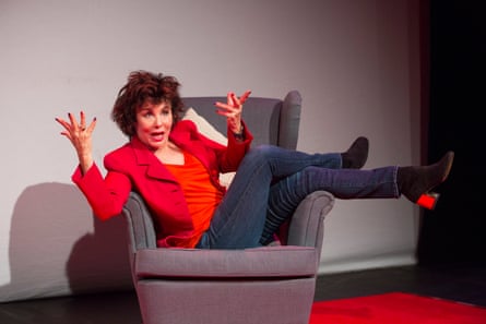 Ruby Wax seated in an armchair onstage, with her legs on the armrest and her hands thrown up