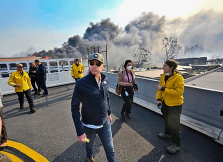 Gavin Newsom walking on a pavement wearing a fleece, jeans, a cap and a pair of shades, with a photographer and a few firefighters around him and smoke billowing in the background