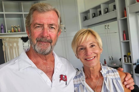 An older white man stands with his arm around the shoulder of an older white woman, both smiling, in a white kitchen. He has a bushy heard and hair, and she has short blond hair.