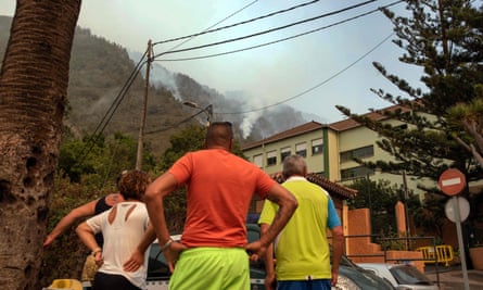People stand in a street looking upwards at columns of smoke from a wildfire burning on forested slopes