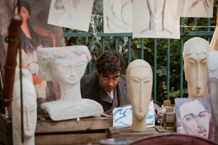 Riccardo Scamarcio sits behind a table holding sculpted heads, with drawings hanging above