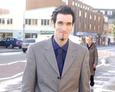 Lewis in a grey suit and blue shirt stands on a street with buildings and cars behind him