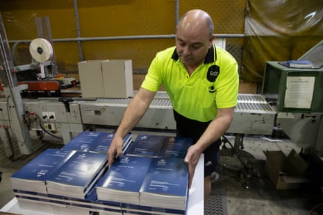 A man wearing a high-vis shirt is stacking budget books on a table in front of a printing and binding machine