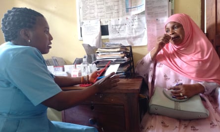 An African nurse seated at a desk and holding tablets talks to an older woman in a hijab