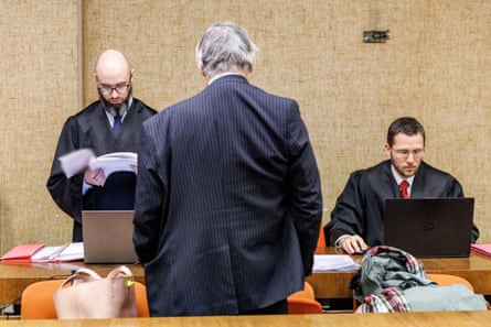 A man in a suit faces away from the camera, looking towards two men in court robes examining files and a laptop.