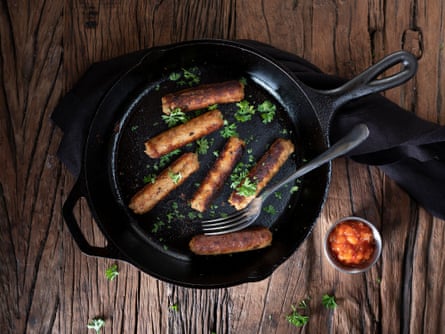 Soya and plant-based vegan sausages in a black skillet on a rustic wooden table; a small container of tomato salsa is to the side, and the sausages are garnished with green parsley.