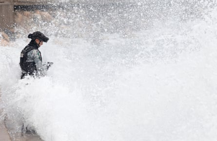man in military uniform holding gun stands by water as ocean water sprays on him
