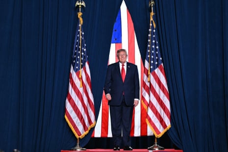 Donald Trump, wearing a blue suit and red tie, stands on stage between blue curtains flanked by american flags