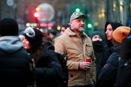he stands among protesters who are mostly dressed in black anoraks but himself wears a light brown jacket and baseball cap; he is tall and in his late 40s with a greyish-red beard