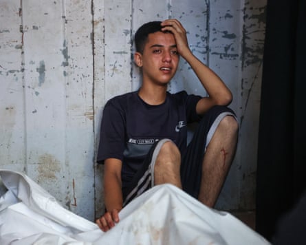 A Palestinian boy sits against a wall crying by a body in a sheet