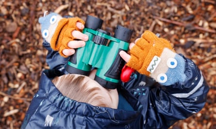 An overhead shot of a young boy standing on wood chip wearing orange fingerless gloves looking through a pair of plastic binoculars