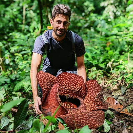 A man in his 30s stands in a jungle environment behind a huge mottled red flower made up of several leaves with a central bowl.