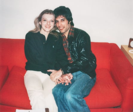 A blond woman in a black top and white jeans, with a dark-haired man in a black leather jacket and jeans, sitting on a red sofa