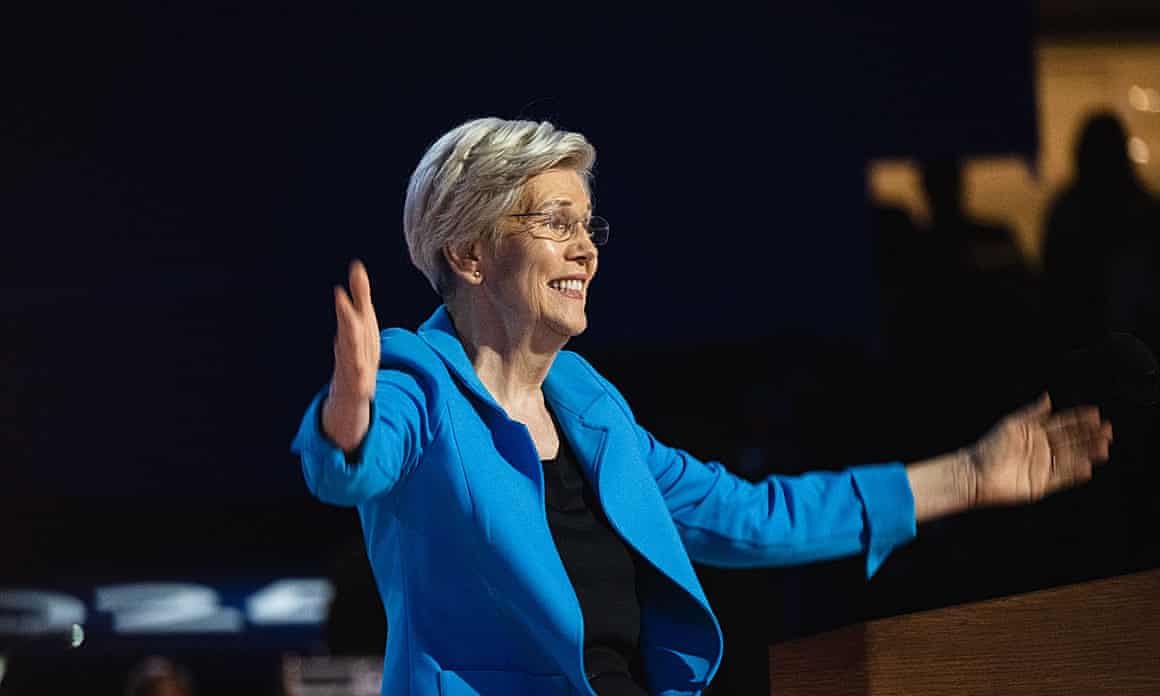 Elizabeth Warren, wearing a light blue jacket, holds her arms out while on stage