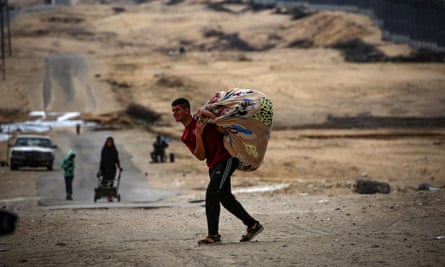 A person carries their belongings, wrapped in a large piece of a material, across a dirt road as another person in the background pulls a handcart along the road