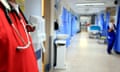 A general view of a hospital corridor with the torso of a medic wearing red scrubs and a stethoscope on the left.