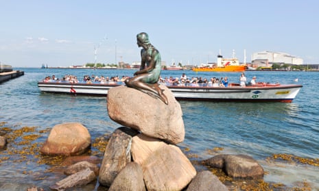 The Little Mermaid statue in the port of Copenhagen, Denmark, with a boat of tourists passing behind.