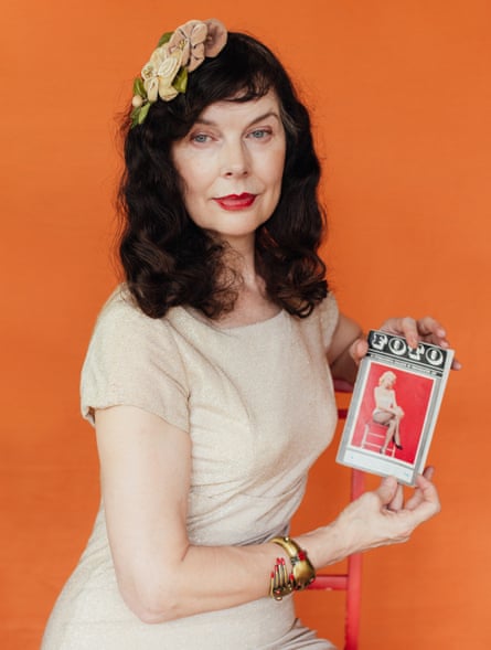 Annastasia sitting at an angle to the camera on a pink chair against an orange wall, wearing a short-sleeved cream dress, a cream and green fascinator, and a gold and red bracelet on her right arm, holding a book called Foto with a woman sitting on a chair on the cover
