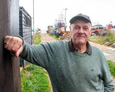 An older man wearing a cap and knitted jumper leans against a black hut. Behind him a path leads past fishing boats to the sea