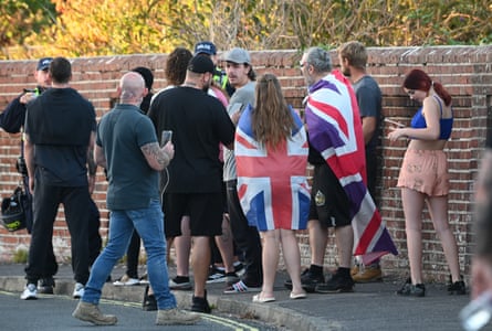 Anti-migrant protesters, some wearing union jacks as cloaks, gather by a wall in front of two police officers in hi-vis jackets.