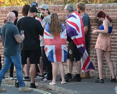 Anti-migrant protesters, some wearing union jacks as cloaks, gather by a wall in front of two police officers in hi-vis jackets.