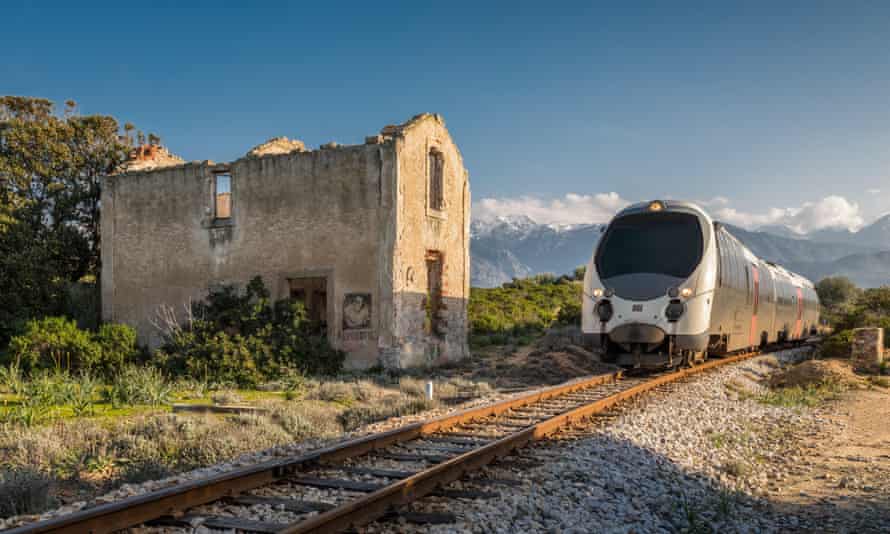 Train passing a derelict station at Lumio, Corsica, with snow-covered mountains in the distance under a deep blue sky.