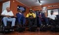 Five Black men sit shoulder to shoulder in barber chairs with orange walls beyond them and a sign that says 'Black Americans for Trump.'