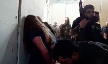 Still from the video showing women lined up facing a wall and soldiers carrying guns.