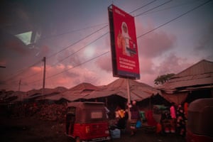 A tuk-tuk passes a row of huts, with an advertising billboard overhead