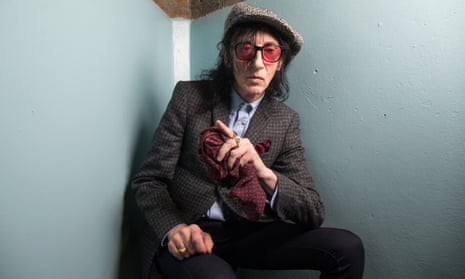 John Cooper Clarke, wearing glasses and a tweed flat cap, sitting in a corner, resting his elbow on his knee