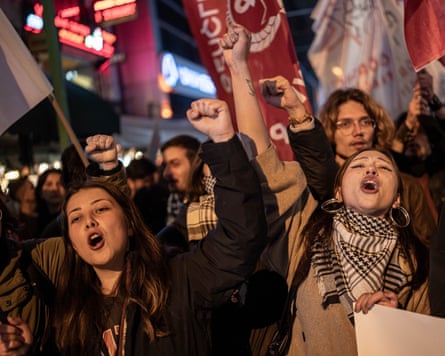 Two women, one with a scarf around her neck, shout at the front of a protest at night with banners behind them, both holding their fists in the air