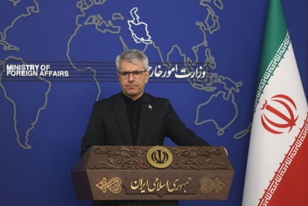 Esmaeil Baghaei speaks at a press conference. He stands against a blue background with an outline map of the region and next to a green, white and red Iranian flag.