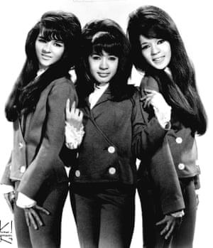 Three women with 60s style hairdos stand together in a black and white photo